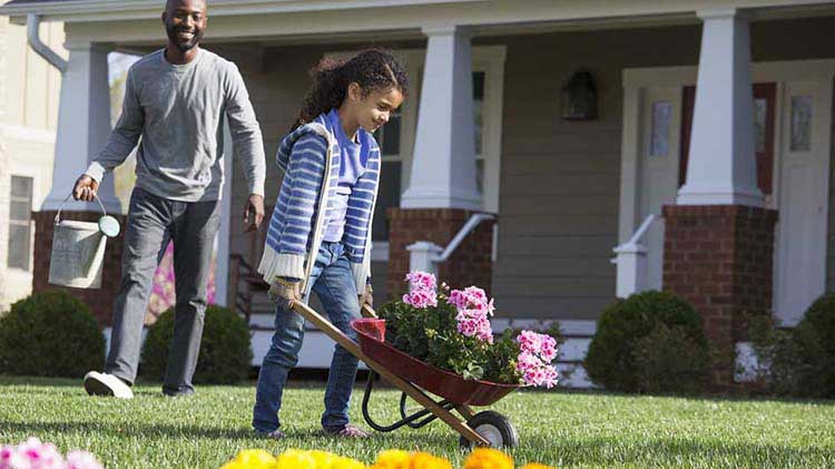 Papá sonriendo a su joven hija que empuja una carretilla con flores para plantar.