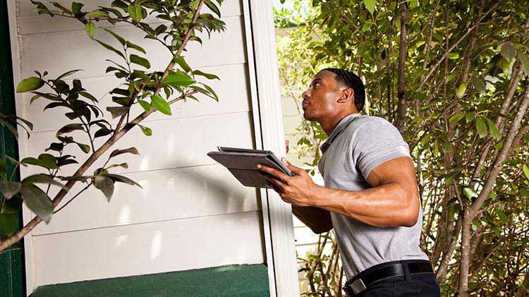 Un hombre mira hacia arriba y sostiene una tableta mientras inspecciona el exterior de una vivienda.