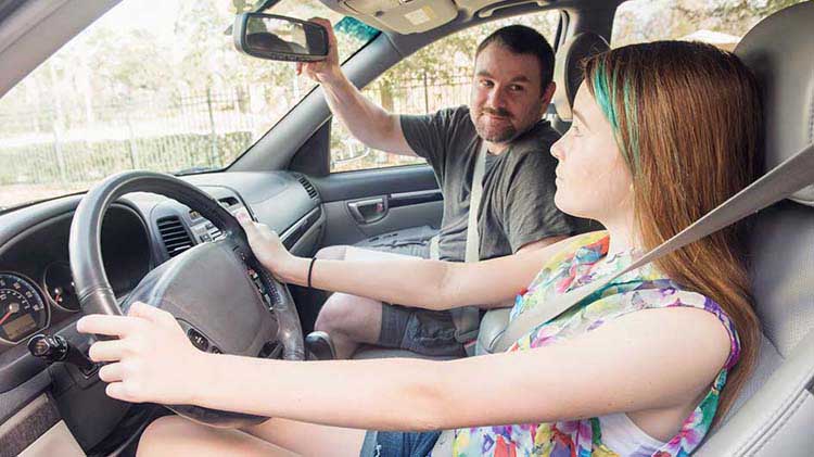 Una adolescente en el asiento del conductor de un carro, usando cinturón de seguridad y con las manos en el volante. Su padre está en el asiento del pasajero ajustando el espejo retrovisor. 