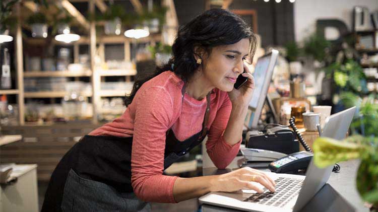 Una mujer propietaria de una pequeña empresa habla por su teléfono celular, en su floristería, mientras escribe en su computadora portátil.