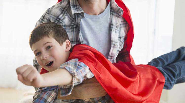 Niño con una capa "volando" en los brazos de su padre.