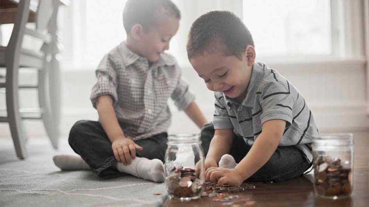 Dos niños sentados en el suelo meten monedas en unos frascos.