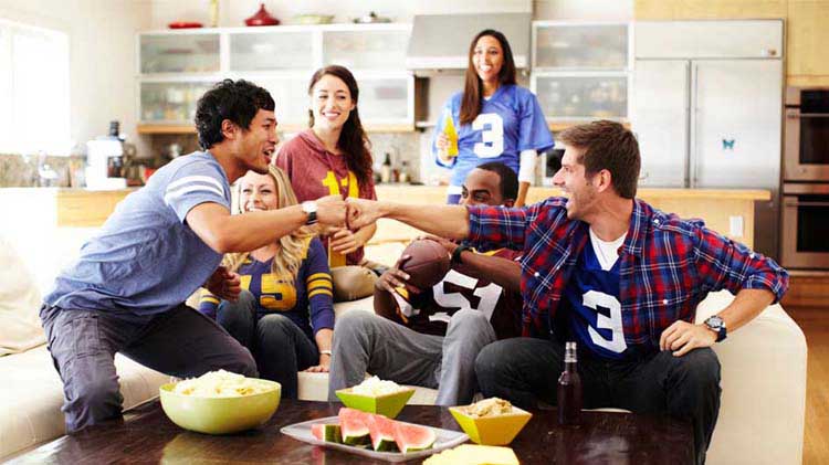 Aficionados de fútbol americano viendo el gran juego y chocando puños mientras consumen bocadillos sentados en un sofá.