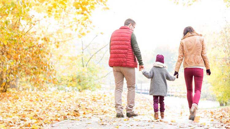 Padres tomados de la mano con su hijo caminando por un sendero cubierto por hojas en el otoño.