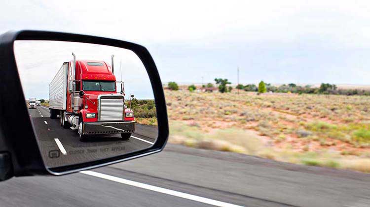 El reflejo de un camión rojo con remolque en el espejo retrovisor de un vehículo.