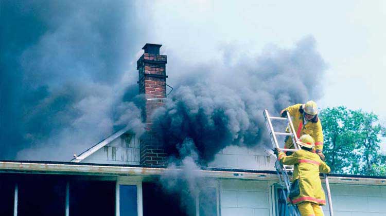 Bomberos a lo alto de una escalera, preparados para apagar un incendio en una casa, mientras humo oscuro sale por el techo.
