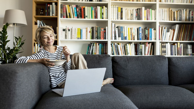 Mujer madura usando un computador portátil para trabajar en el plan del patrimonio digital y está almorzando en el sillón.