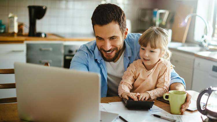 Un hombre con su hija sentada en su regazo en la mesa de la cocina trabajando en su computadora portátil, explorando diferentes tipos de cuentas de ahorros.