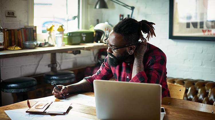 Hombre barbudo con gafas en el escritorio de la casa con computadora portátil, revisando un documento en papel.