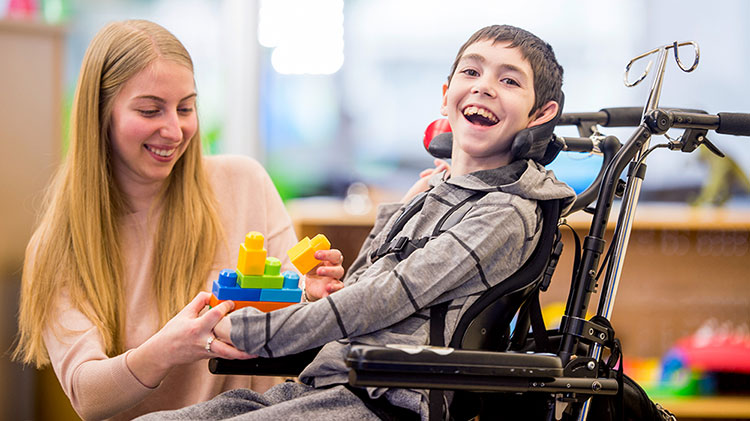 Un niño en silla de ruedas sonríe mientras una mujer le entrega bloques de juguetes.