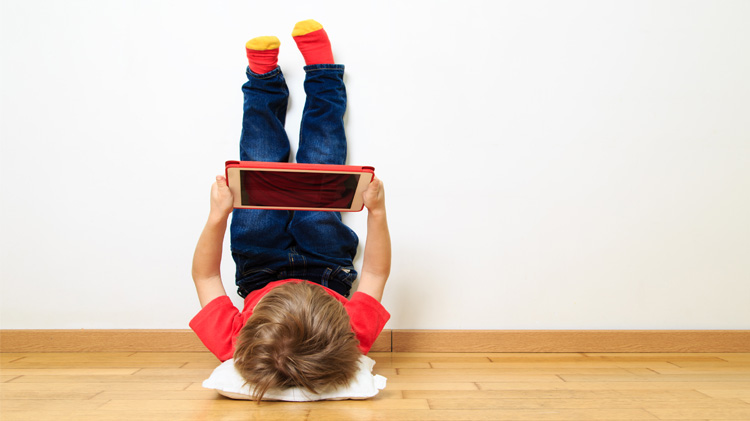 Niño pequeño acostado en un piso de madera jugando en su tableta con los pies apoyados en la pared.