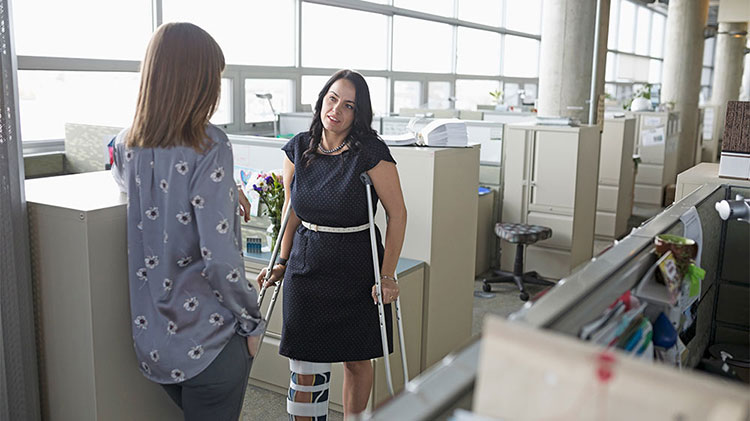 Dos mujeres de pie en la oficina y una tiene un aparato ortopédico en la pierna y está usando muletas.
