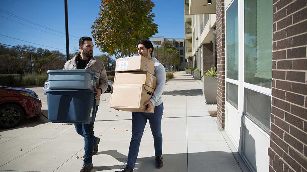 Dos personas sosteniendo cajas y contenedores de mudanza, caminando por una acera frente a un edificio de apartamentos.