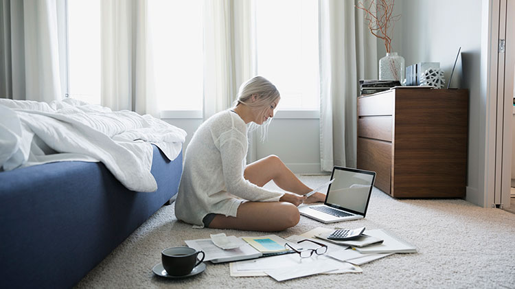 Una mujer sentada en el suelo frente a su computadora y revisando cuentas.