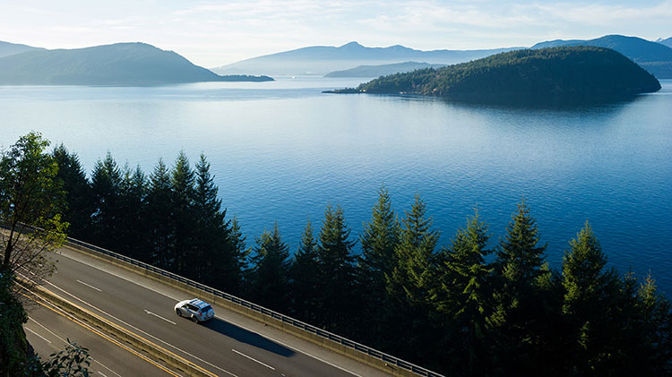 Un carro circula por la autopista junto a un lago y unas montañas