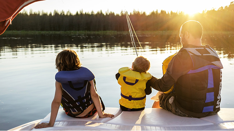 Un padre y dos niños sentados en una embarcación pescan en el lago, en un día soleado.